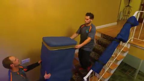 two uniformed movers carrying a blanket-wrapped sofa down a staircase, face visible